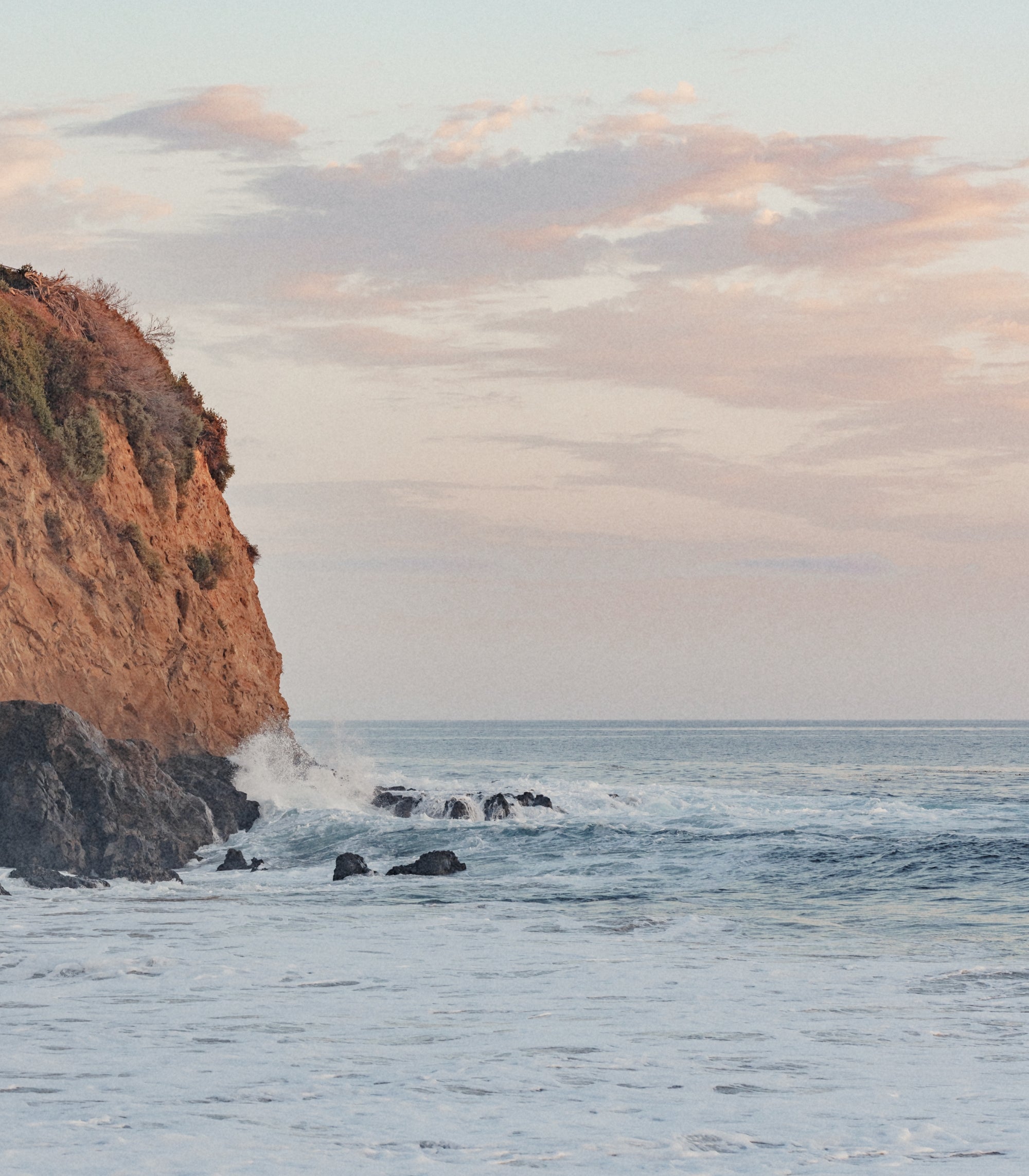 A coastal seascape with a rocky cliff on the left, ocean water with small waves, dark rocks in the surf, and a pale sky with soft pink clouds.