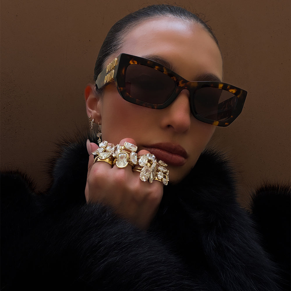 Editorial fashion portrait of a model wearing an extravagant assortment of sculptural yellow gold diamond statement rings featuring oval, marquise, pear, and elongated diamond cuts, paired with delicate diamond drop earrings. Styled in oversized tortoiseshell sunglasses and luxurious black fur against a warm brown backdrop for a bold, glamorous holiday fine jewelry look by Sofia Kaman Fine Jewels.