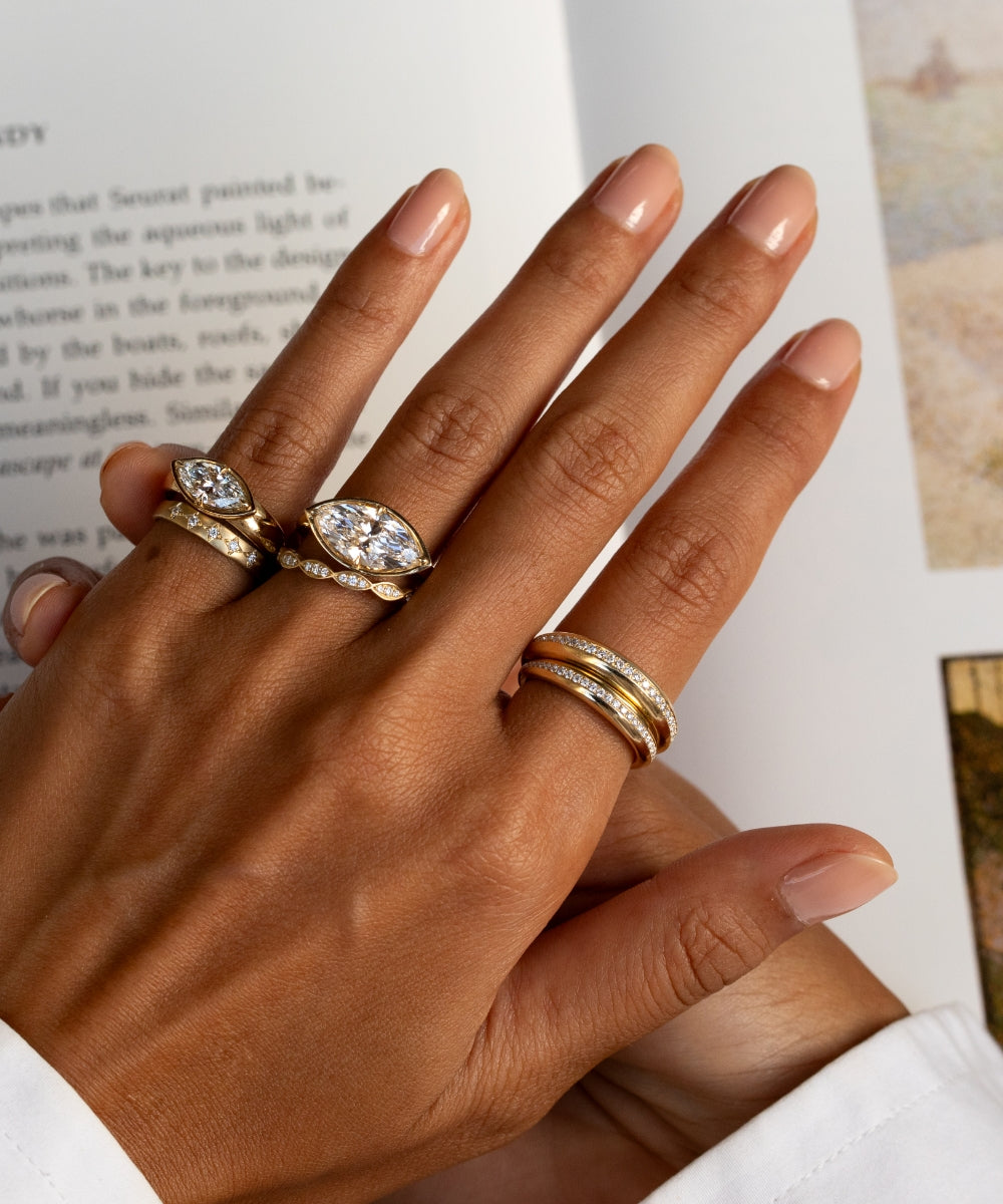 Close-up of a hand with a book in the background, styled with yellow gold and diamond rings, featuring two east-west marquise diamond rings paired with delicate diamond bands and a layered pavé pinky ring stack.