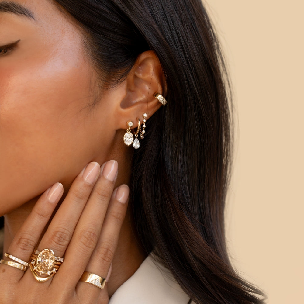 Close-up of a woman wearing a layered mix of gold and diamond earrings, including oval and pear-shaped diamond drops, delicate hoops, and an ear cuff. Her hand is raised near her face, showcasing a stack of sculptural gold and diamond rings.