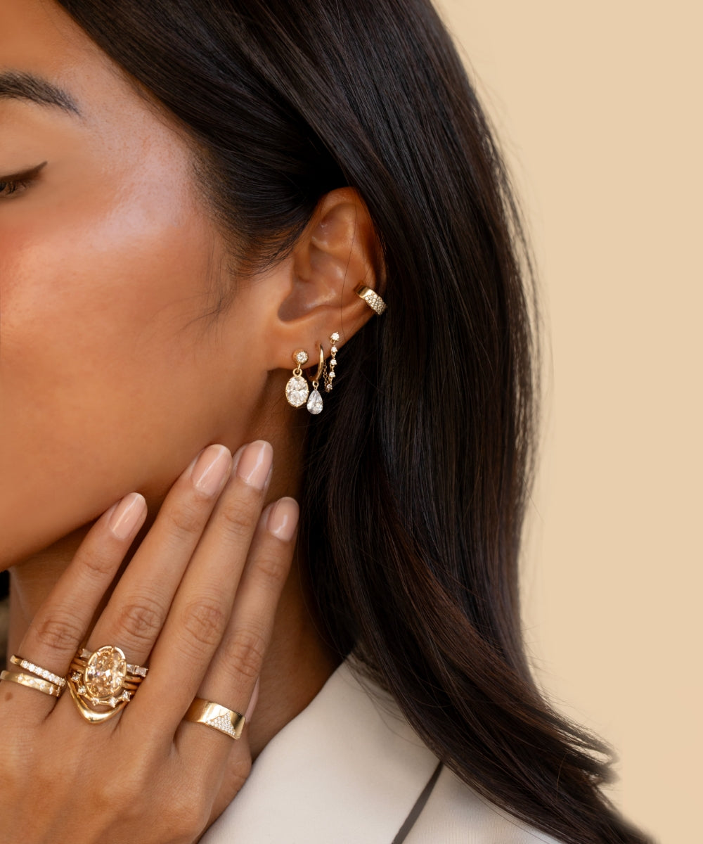 Close-up of a woman wearing a layered mix of gold and diamond earrings, including oval and pear-shaped diamond drops, delicate hoops, and an ear cuff. Her hand is raised near her face, showcasing a stack of sculptural gold and diamond rings.