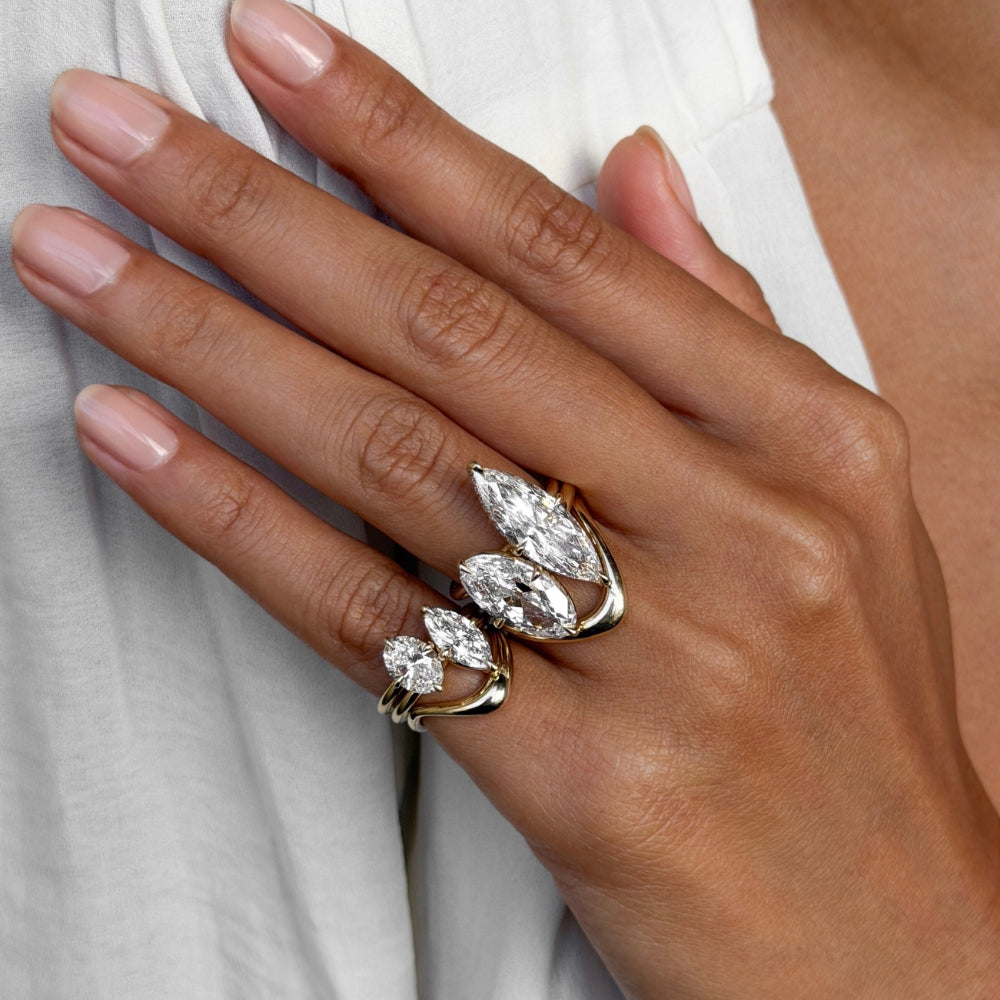 A close-up of a hand wearing a gold ring set with marquise-cut clear gemstones, resting against a white garment.