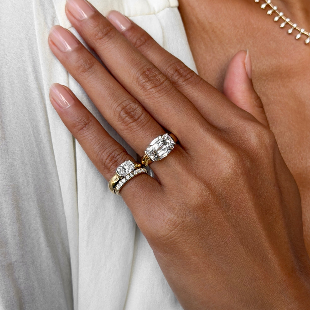 A close-up of a hand wearing two rings: a yellow gold ring with a large oval-cut clear gemstone and a silver-toned band with small clear stones, against a white draped garment.