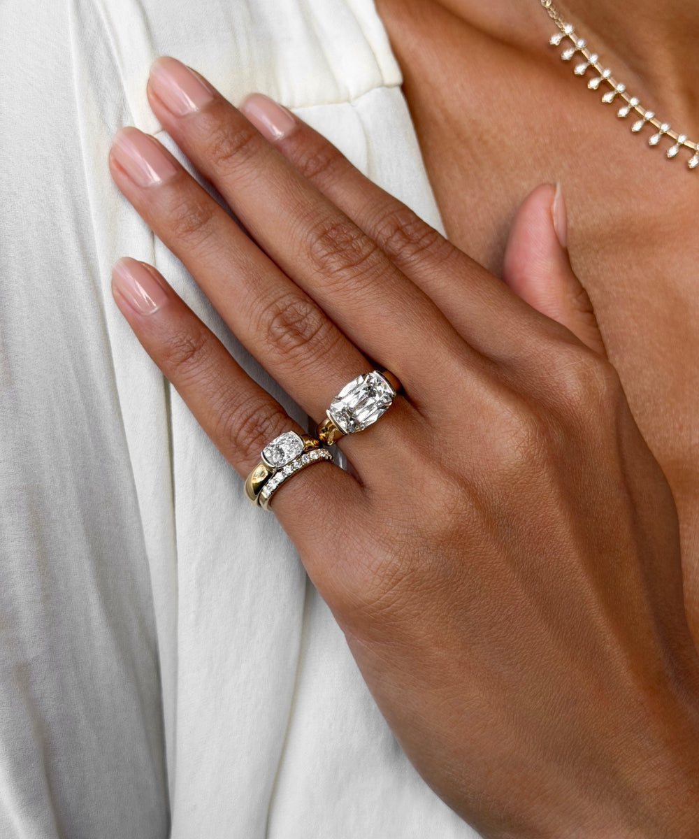 A close-up of a hand wearing two rings: a yellow gold ring with a large oval-cut clear gemstone and a silver-toned band with small clear stones, against a white draped garment.