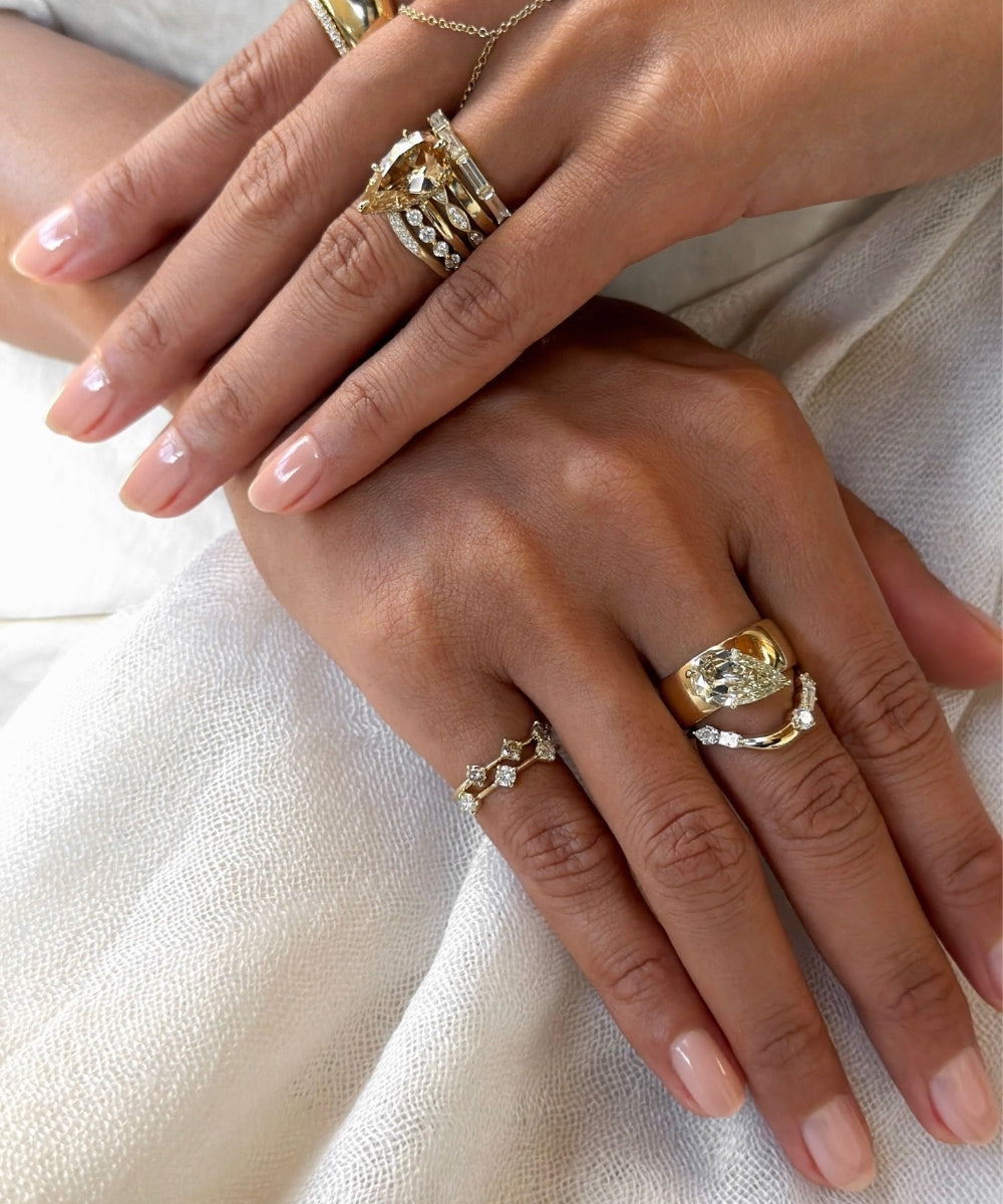 Close-up of two hands styled with layered yellow gold and diamond rings, featuring pear-shaped champagne and white diamonds, sculptural gold bands, and delicate diamond accent pinky rings against soft white fabric.