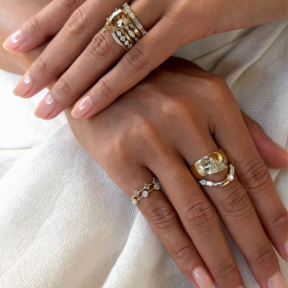 Close-up of two hands styled with layered yellow gold and diamond rings, featuring pear-shaped champagne and white diamond stacking bands, sculptural gold pear shaped band, delicate diamond pinky rings, and a fine gold hand chain.