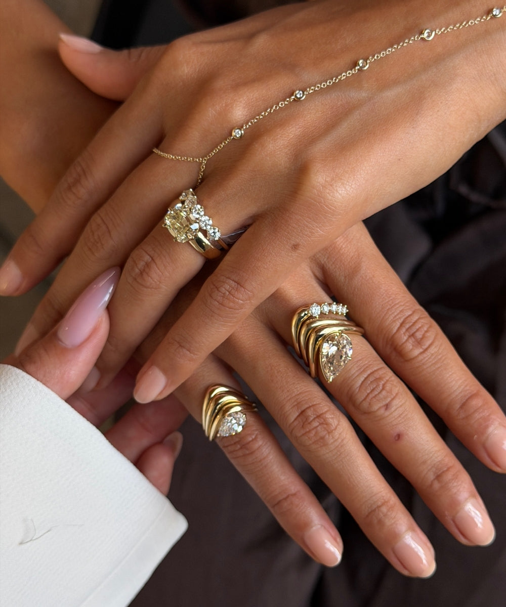 Close-up of two hands layered with sculptural yellow gold and diamond rings, featuring pear and elongated cushion-cut diamonds, delicate diamond accents, and a fine gold hand chain.