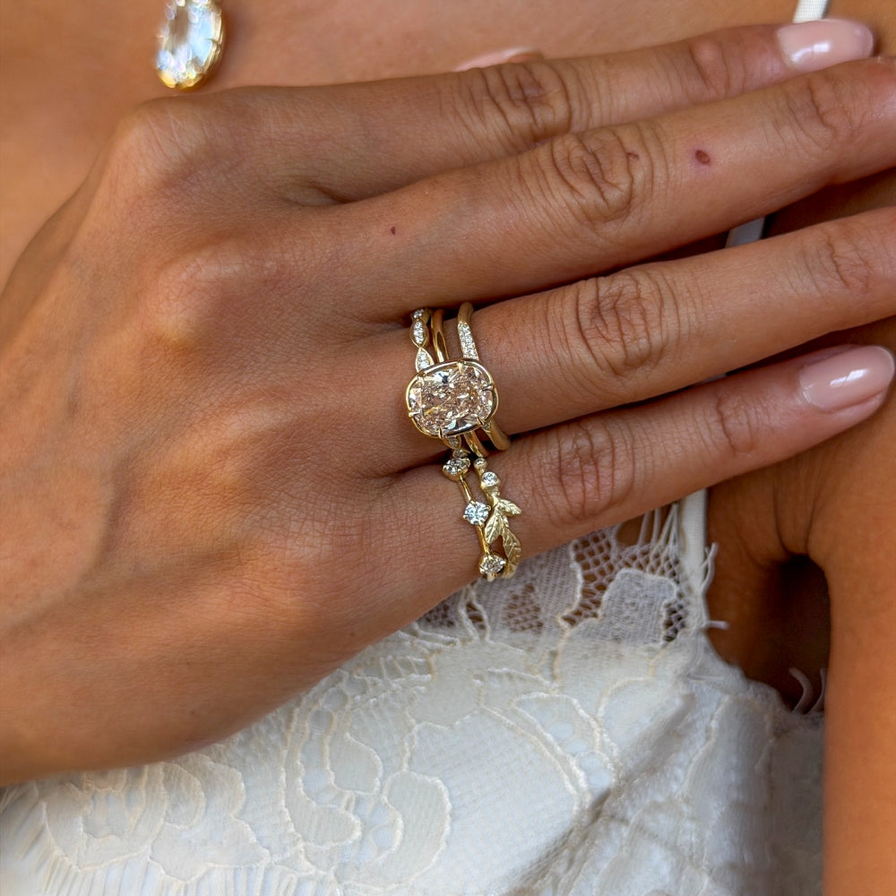 Close-up of a hand wearing a layered stack of yellow gold and diamond rings, centered around an oval champagne diamond engagement ring and accented with delicate pavé and botanical-inspired bands and pinky rings, against a white lace dress.