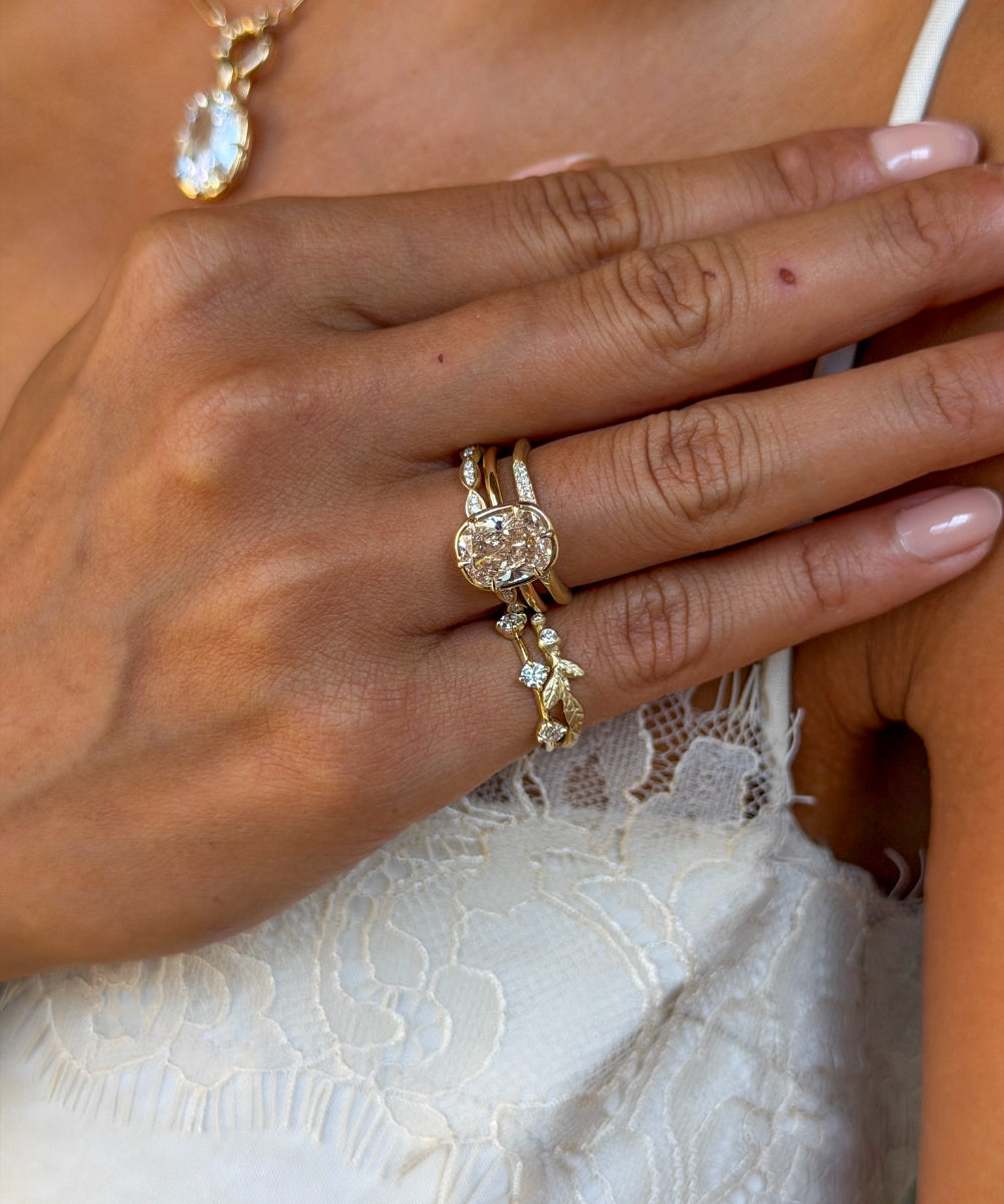 Close-up of a hand wearing a layered stack of yellow gold and diamond rings, centered around an oval champagne diamond engagement ring and accented with delicate pavé and botanical-inspired bands and pinky rings, against a white lace dress.