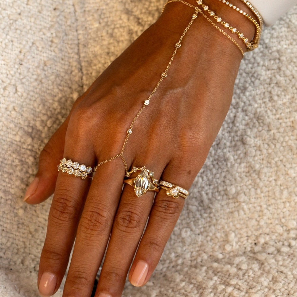 A close-up of a hand wearing a gold chain hand bracelet with delicate links, paired with multiple gold rings and stacked gold bangles on a textured beige fabric background.