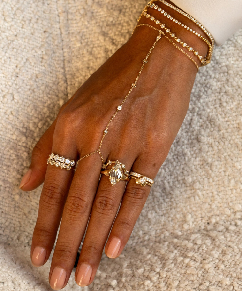 A close-up of a hand wearing a gold chain hand bracelet with delicate links, paired with multiple gold rings and stacked gold bangles on a textured beige fabric background.