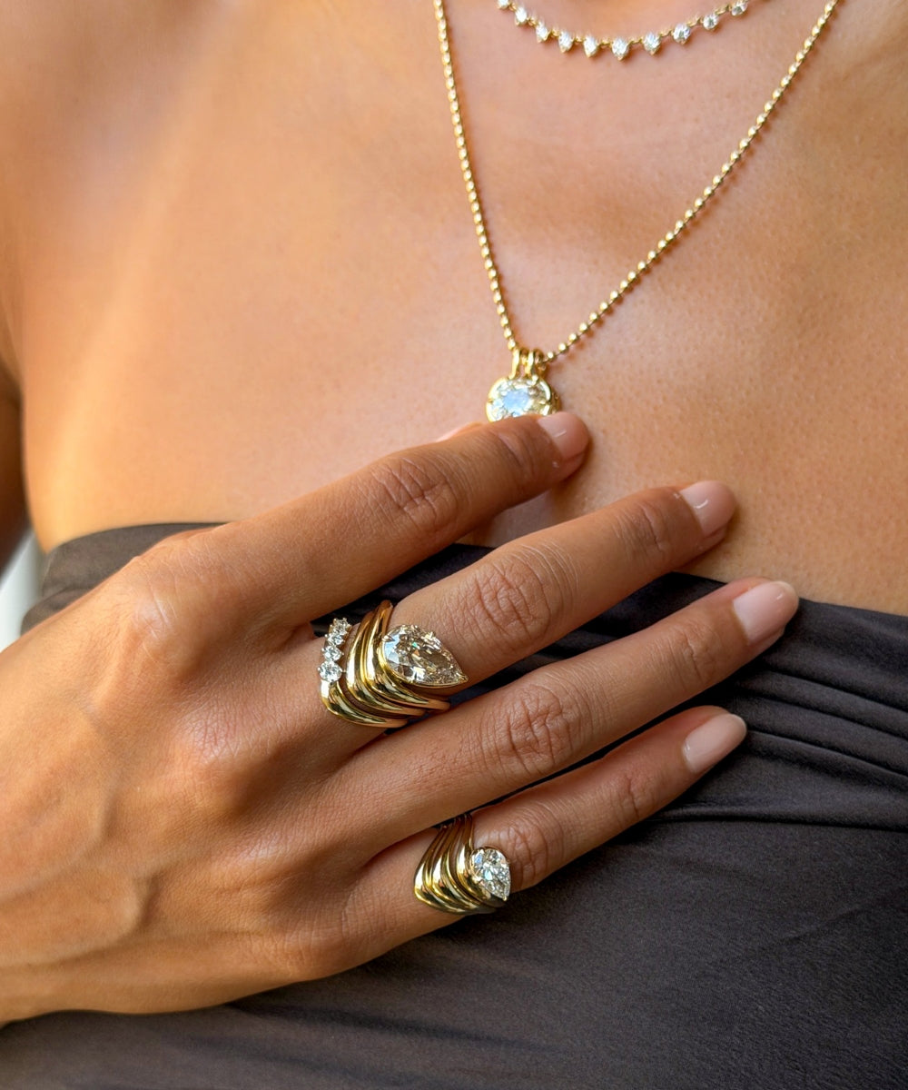Gold rings with clear gemstones worn on a woman's fingers, shown in a close-up lifestyle shot against her chest and dark gray top.