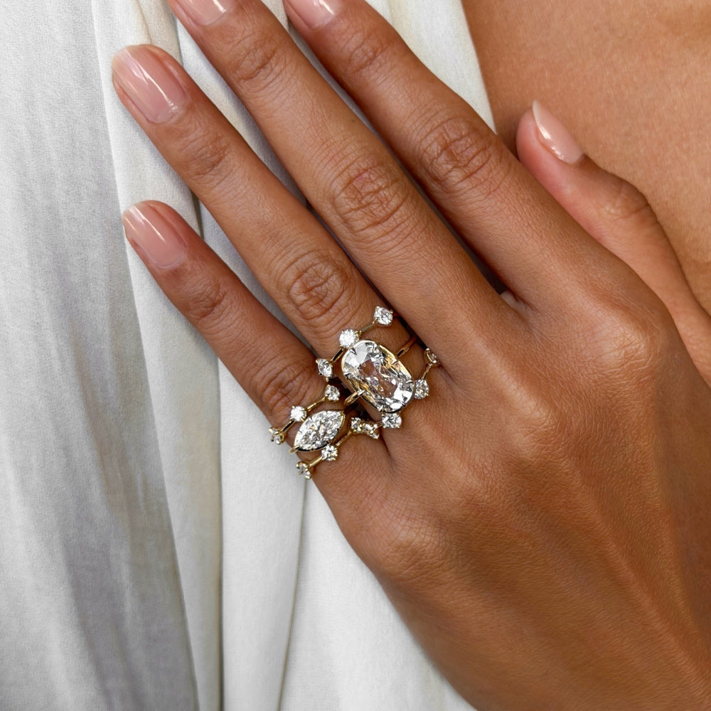 Close-up of a hand wearing layered yellow gold and diamond rings, featuring a large cushion old mine cut diamond and marquise diamond pinky ring surrounded by delicate scattered diamond accent pinky rings, styled against soft white fabric.