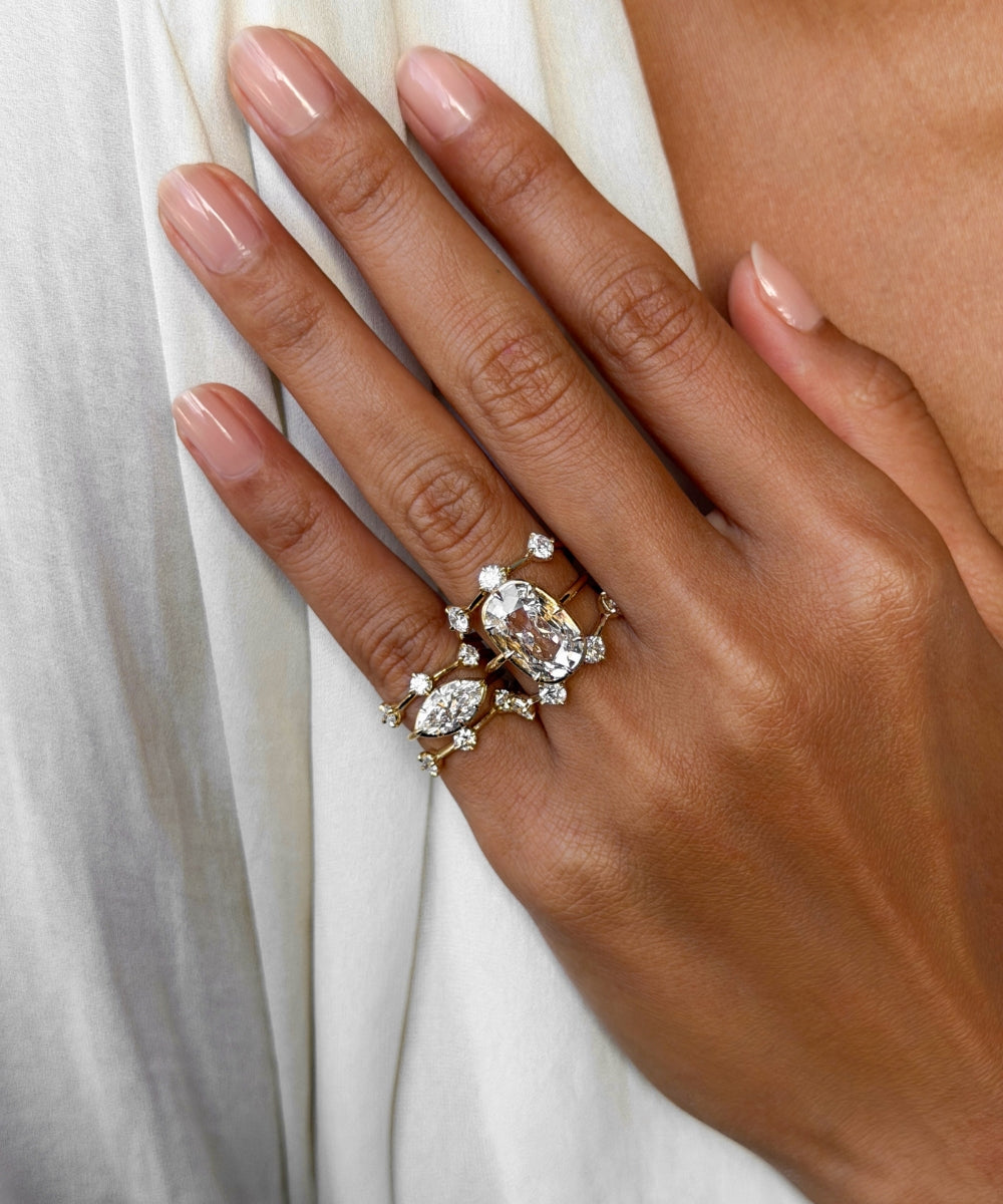 Close-up of a hand wearing layered yellow gold and diamond rings, featuring a large cushion old mine cut diamond and marquise diamond pinky ring surrounded by delicate scattered diamond accent pinky rings, styled against soft white fabric.