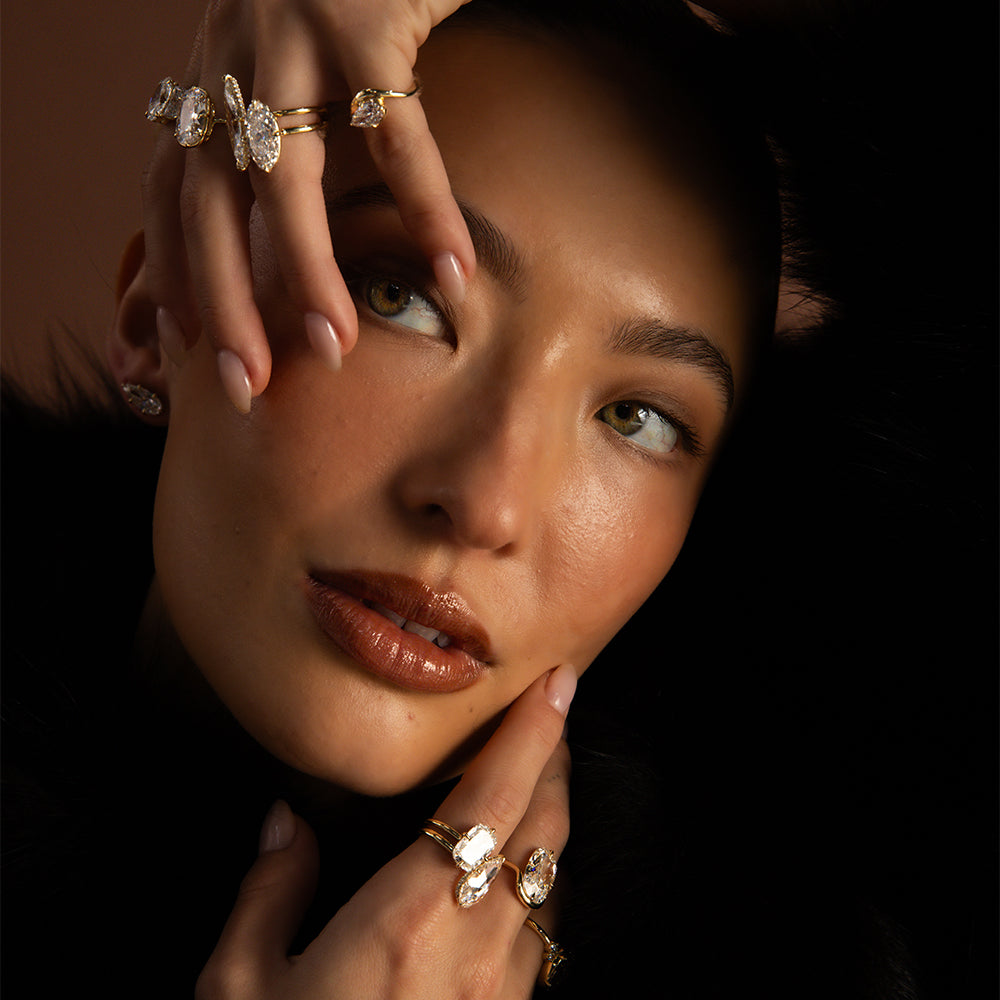 Editorial beauty portrait of a model wearing sculptural 14K yellow gold diamond rings featuring marquise, oval, and pear-shaped diamonds, paired with sparkling Annika lab diamond stud earrings by Sofia Kaman Fine Jewels. Hands elegantly frame her face, showcasing layered statement rings against a dramatic black fur backdrop with warm, moody lighting for a luxurious holiday fine jewelry look.