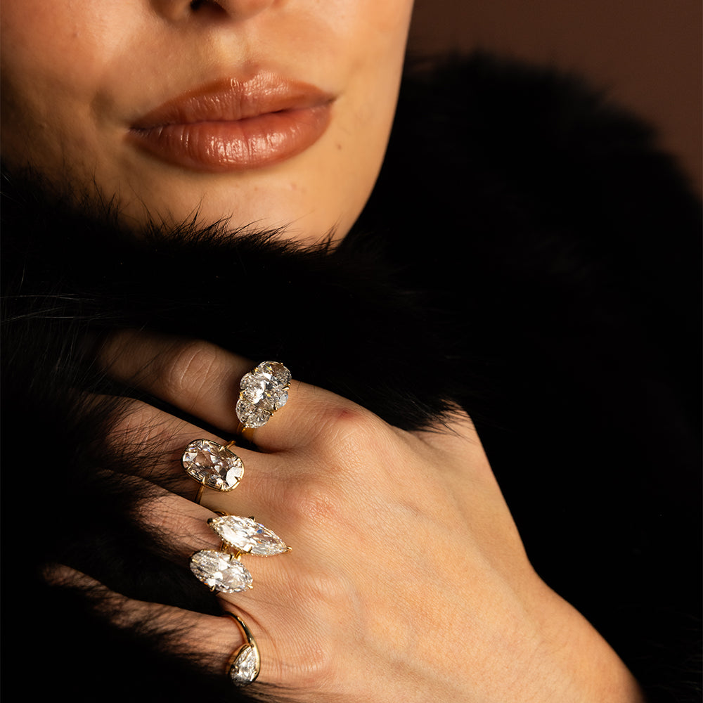 Close-up editorial photograph of a model wearing an eclectic stack of Sofia Kaman 14K yellow gold diamond rings, featuring sparkling oval, marquise, pear, and antique-inspired diamond cuts. Her hand rests against a luxurious black fur collar, with warm lighting highlighting the sculptural gold settings, brilliant diamonds, and bold, modern fine jewelry styling.