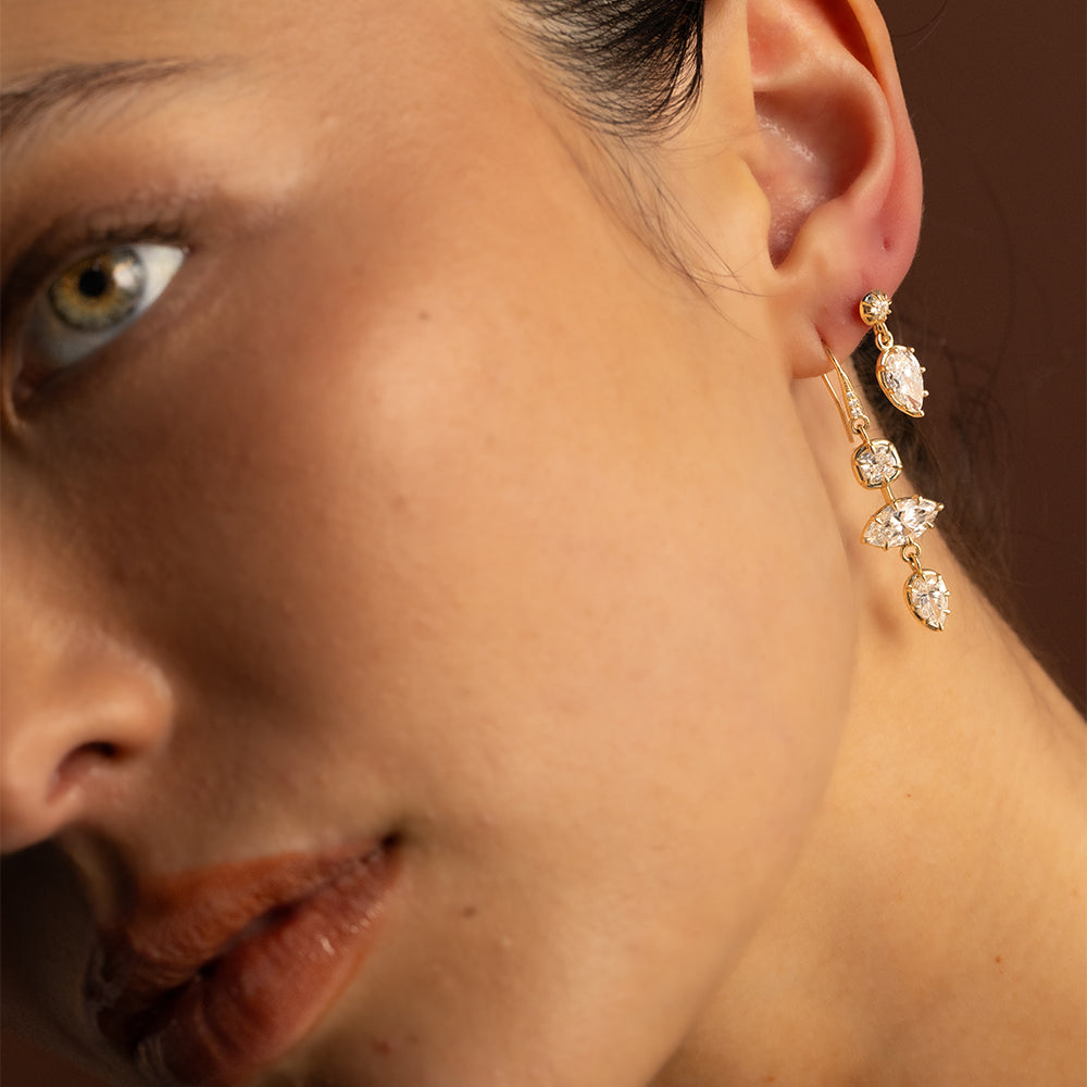Editorial close-up of a model wearing layered 14K yellow gold diamond drop earrings by Sofia Kaman Fine Jewels, featuring sparkling pear, marquise, and cushion-shaped diamonds in delicate dangling settings. Warm, soft lighting highlights the intricate gold details and brilliant diamonds for a modern, sophisticated fine jewelry look.