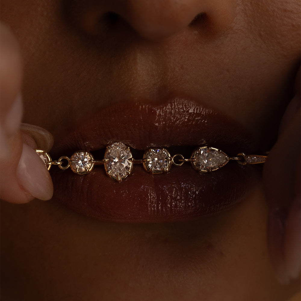 Close-up editorial photograph of a model holding a delicate 14K yellow gold diamond jewelry gigi duster across her lips, featuring round, oval, cushion, and pear-shaped diamonds in vintage-inspired settings. Deep, moody lighting highlights the sparkling diamonds against glossy lips, creating a sensual, modern fine jewelry look for Sofia Kaman Fine Jewels.