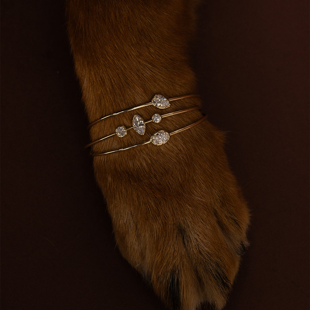 Editorial close-up of three sculptural 14K yellow gold Gigi diamond cuff bracelets stacked around a rich brown fur-covered Rotteweiler paw, featuring pear, marquise, and round brilliant cut diamonds. Styled against a dark chocolate brown background for a playful, luxurious holiday fine jewelry look by Sofia Kaman Fine Jewels.