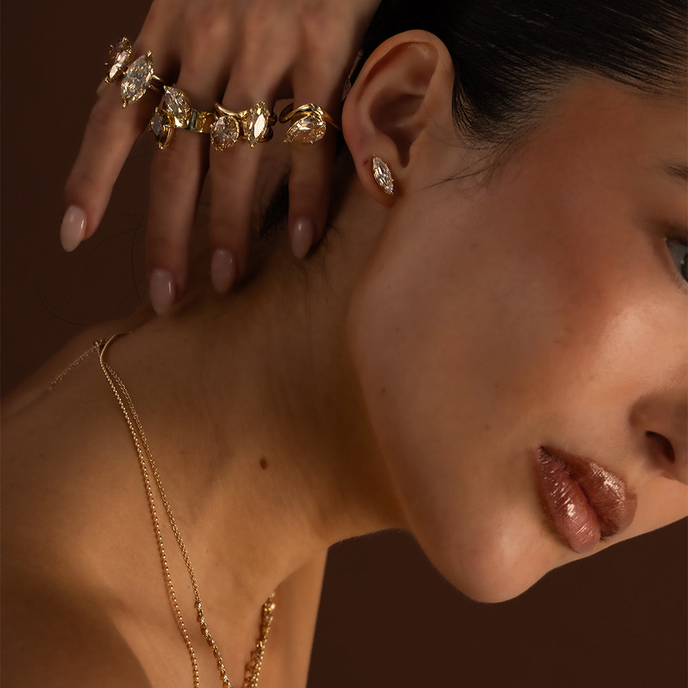 Editorial beauty close-up of a model wearing an assortment of sculptural yellow gold diamond rings featuring marquise, pear, and oval-shaped diamonds, paired with a Annika marquise lab diamond stud earring and layered delicate gold necklaces. Styled with sleek dark hair and warm, moody lighting against a rich chocolate brown background for a sophisticated holiday fine jewelry look by Sofia Kaman Fine Jewels.