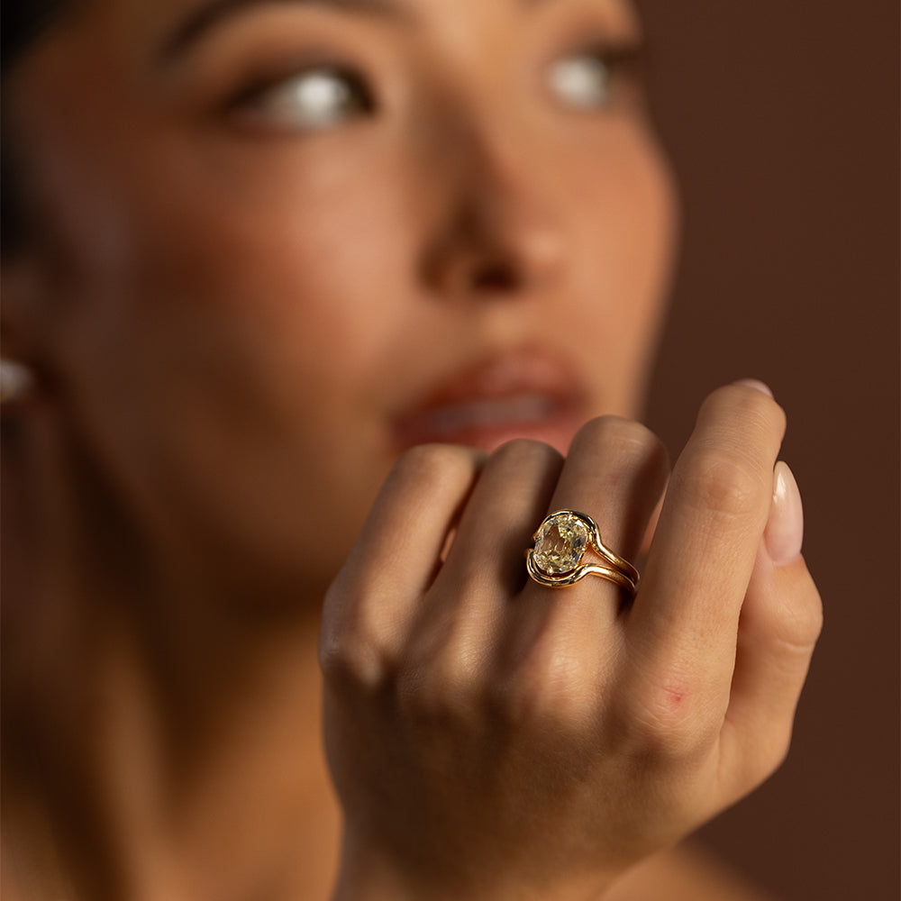 Close-up editorial photograph of a model wearing a Sofia Kaman marquise-cut champagne diamond ring in a sculptural yellow gold setting. Her hand rests gently against her lips, highlighting the elongated diamond, warm golden tones, and elegant silhouette against a softly blurred, moody brown background.