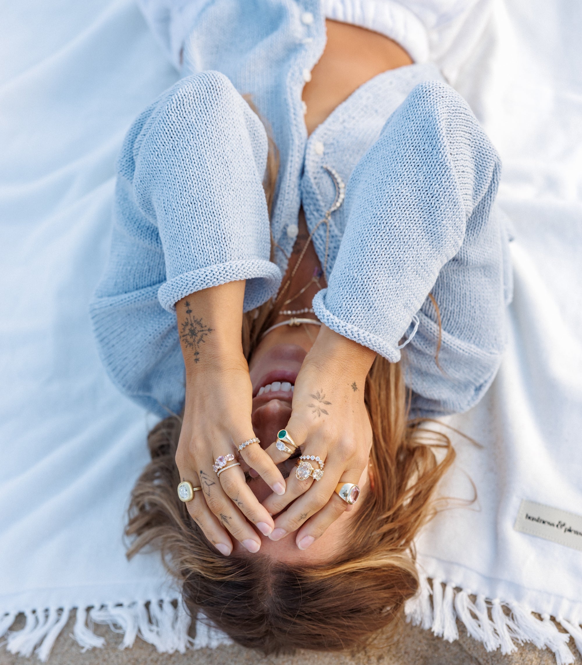Julia Havens lying on a white fringed blanket, wearing a light blue knit cardigan and multiple silver rings with colorful gemstones.