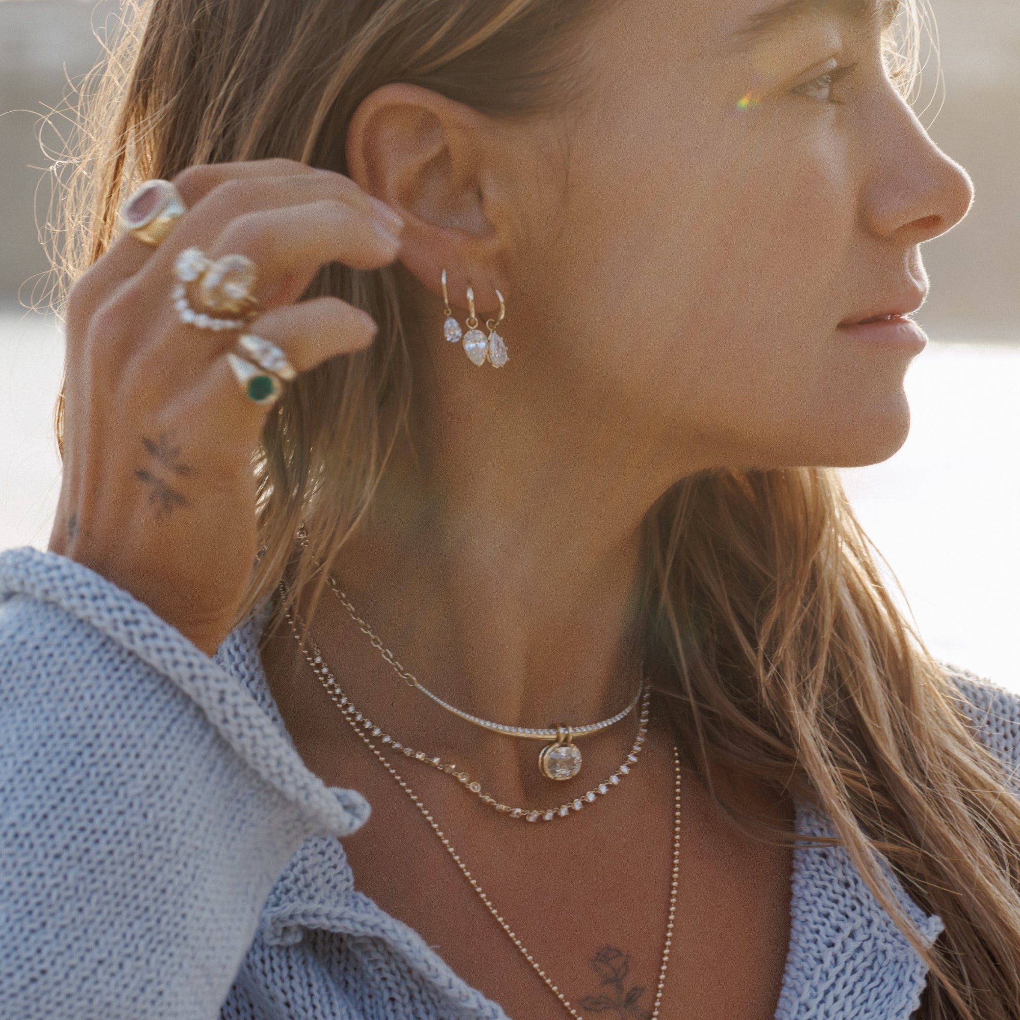 Brunette Woman On Beach With Sun Behind Her Showing Off Gorgeous Ring And Earring Stack Of Solid Gold Fine Jewelry