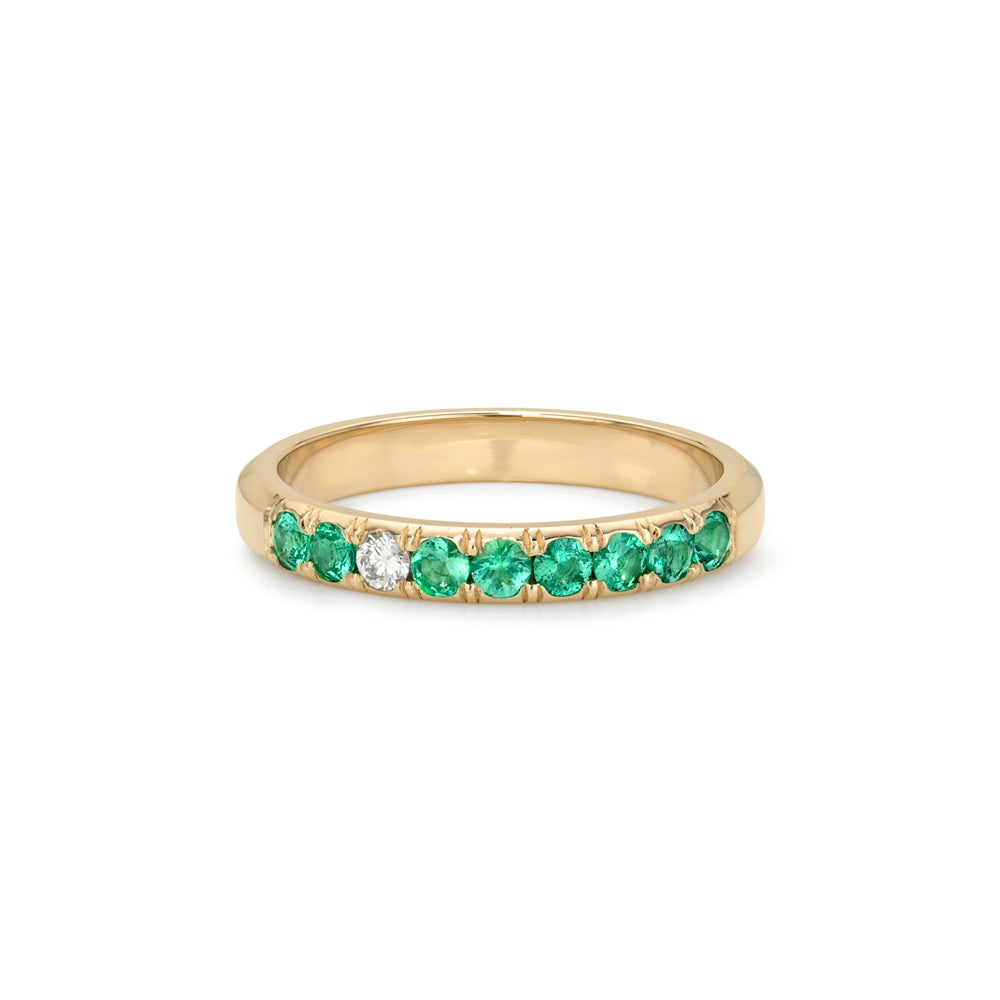 A yellow gold eternity ring featuring a row of round-cut green emeralds with one central clear diamond, set against a plain white background.