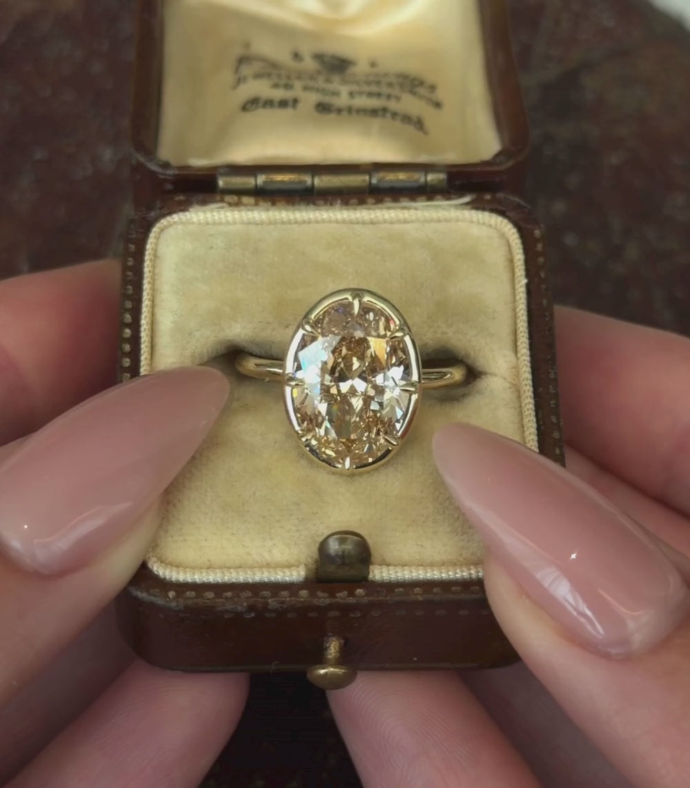 A yellow gold ring featuring a large Oval brilliant cut champagne diamond in a eight-prong setting, shown against a vintage ring box and hands modeling.