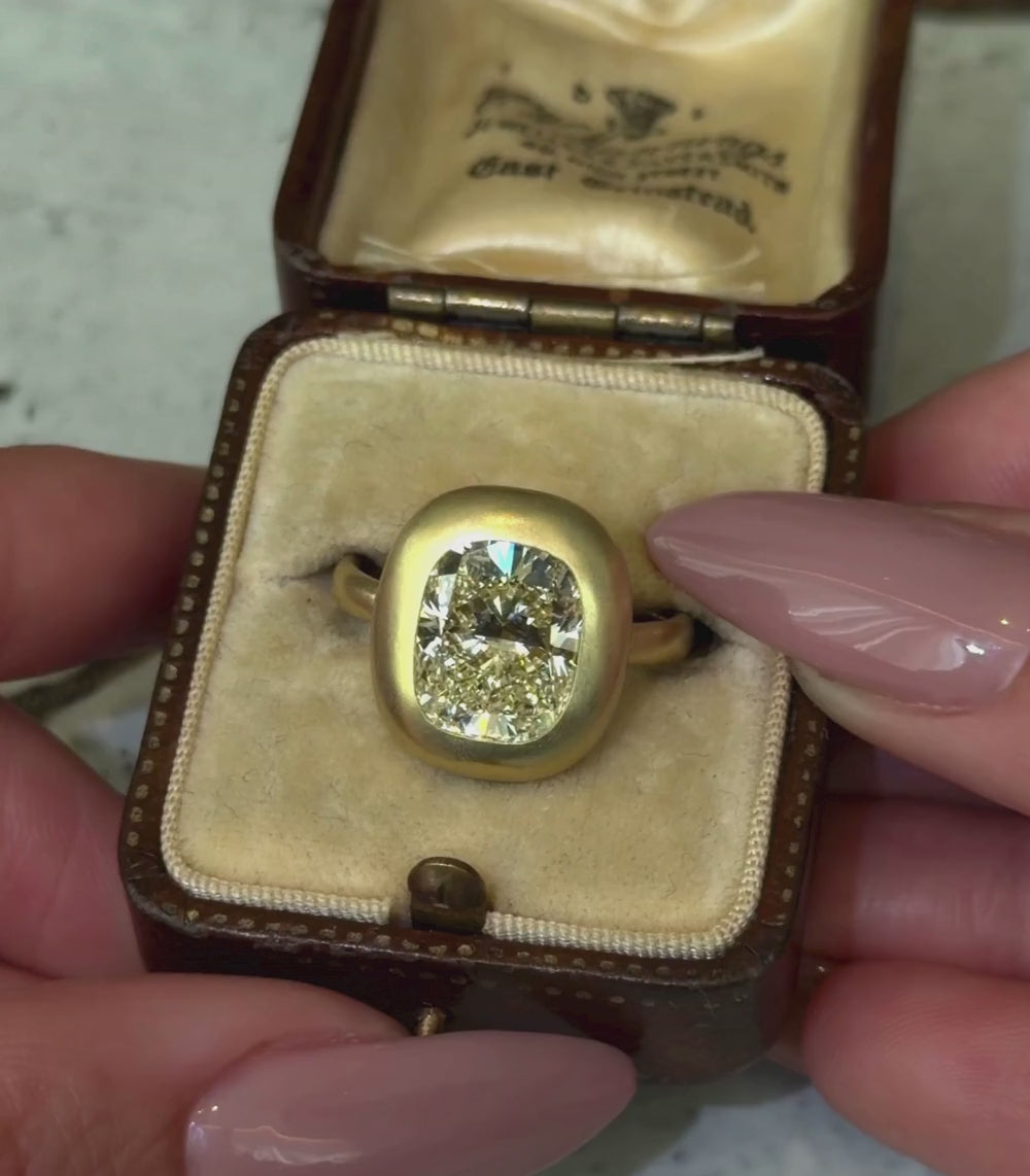 18K yellow gold ring with a 5.02ct elongated cushion brilliant cut champagne diamond bezel set in a engagement ring setting shown in a box with hands.