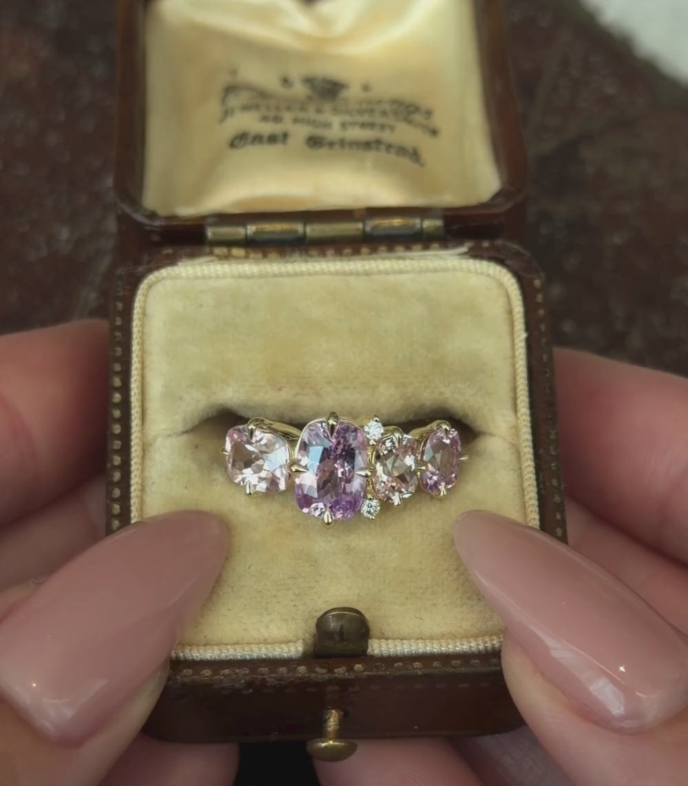 A yellow gold ring featuring several cushion shaped pink and purple sapphires with round brilliant pave stones in cluster prong setting, shown against a vintage ring box and hands modeling.