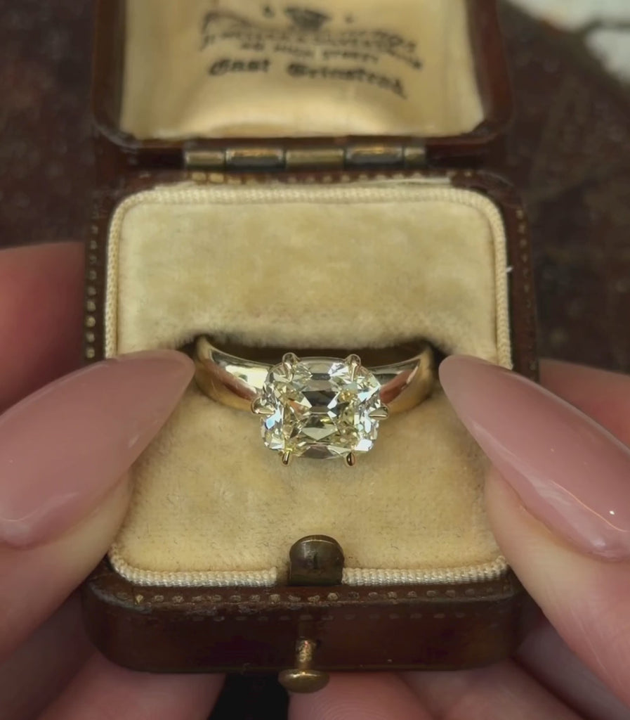 A yellow gold ring featuring a large cushion old mine cut champagne diamond in a six-prong setting, shown against a vintage ring box and hands modeling.