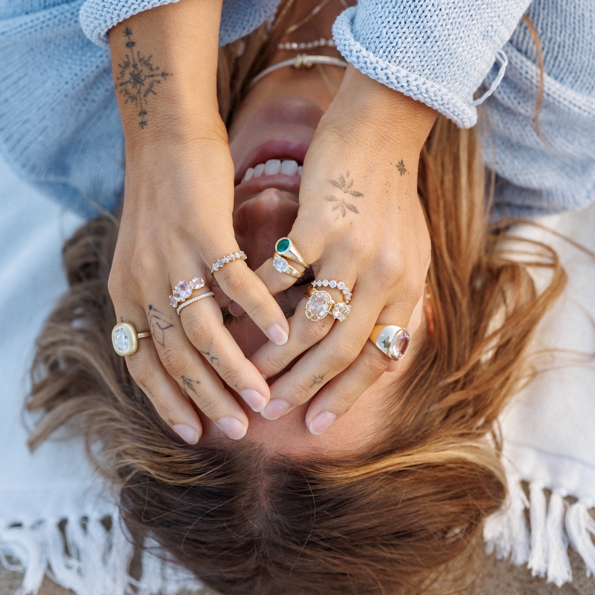 Woman Laying On Beach Towel In Blue Knit Sweater With Hands Ver Face With Various Types Of Diamond And Gemstone Engagement Rings