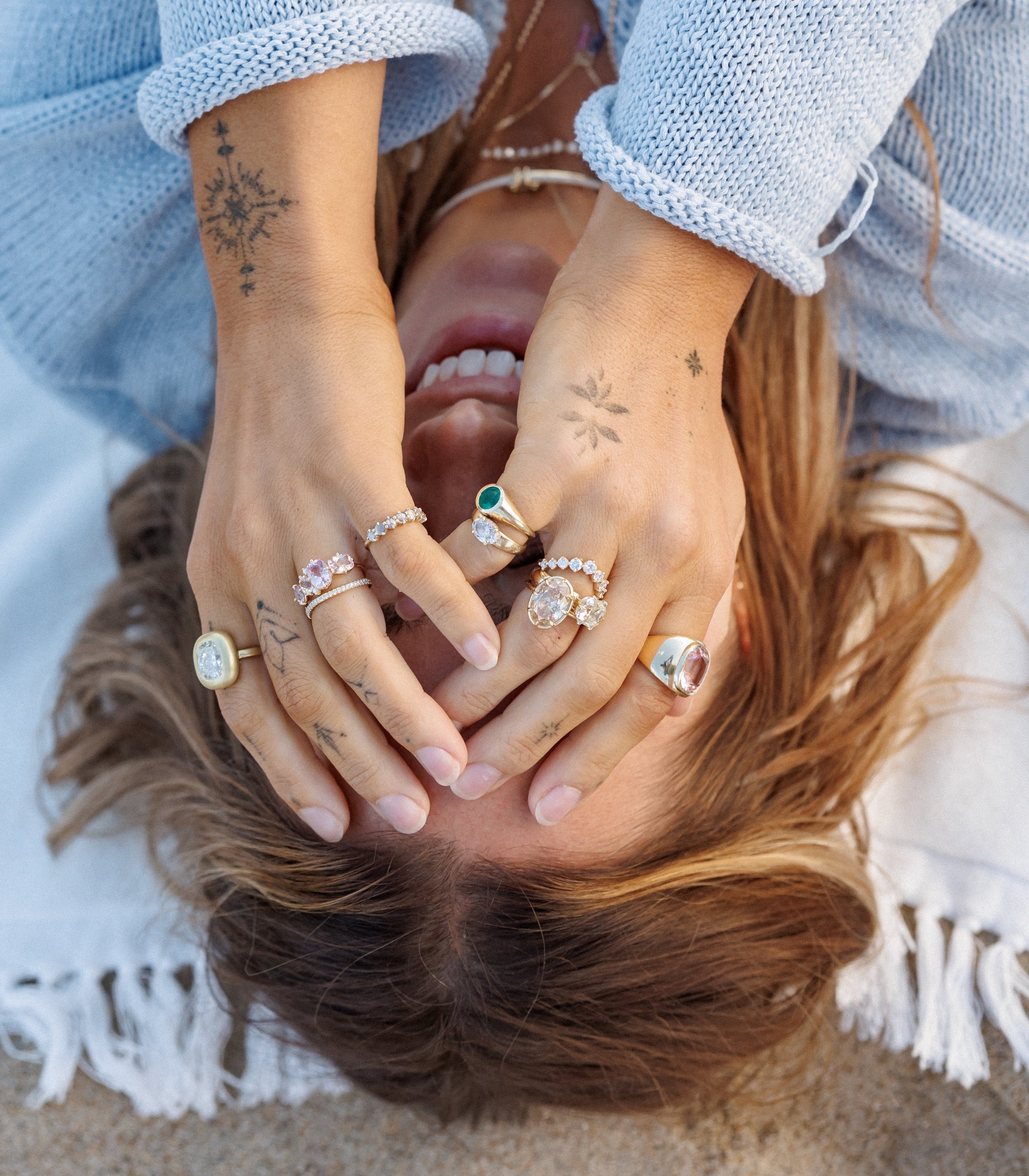 Woman Laying On Beach Towel In Blue Knit Sweater With Hands Ver Face With Various Types Of Diamond And Gemstone Engagement Rings