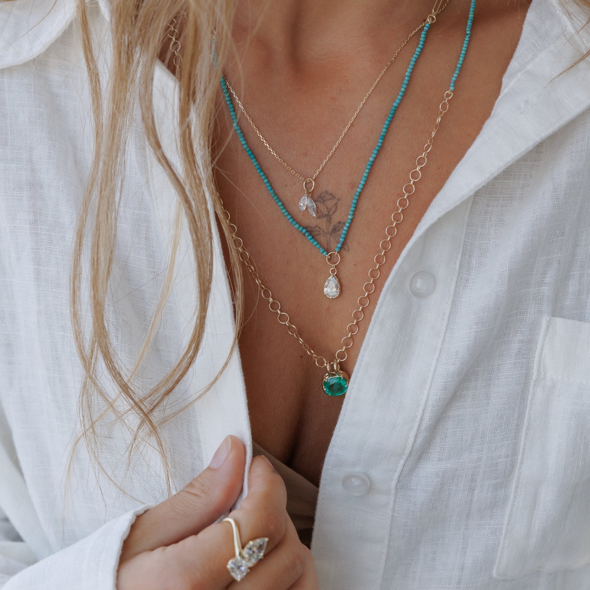 Woman Wearing White Linen Button Down Shirt With Layers Of Necklaces With Diamond And Emerald Pendants