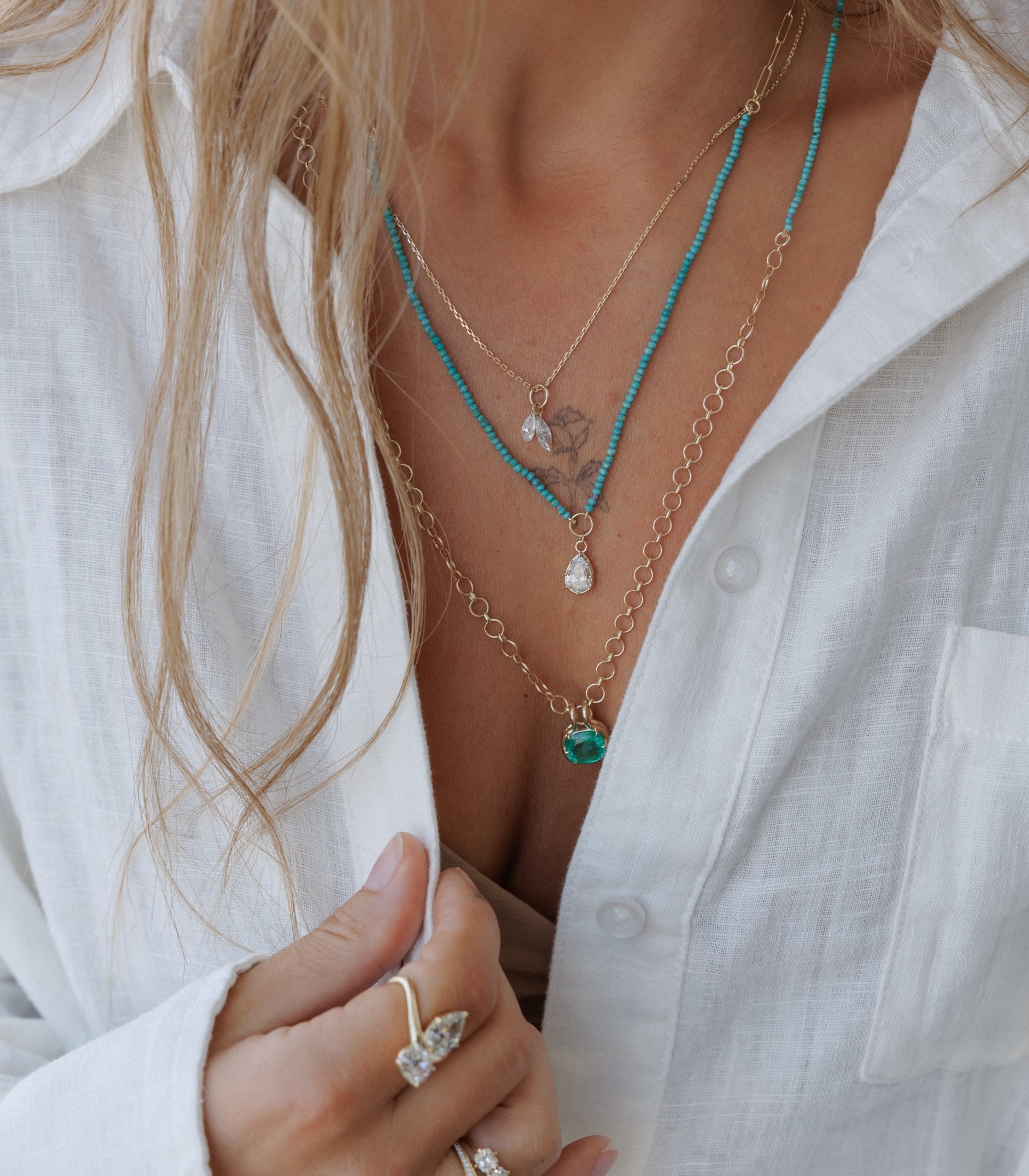 Woman Wearing White Linen Button Down Shirt With Layers Of Necklaces With Diamond And Emerald Pendants