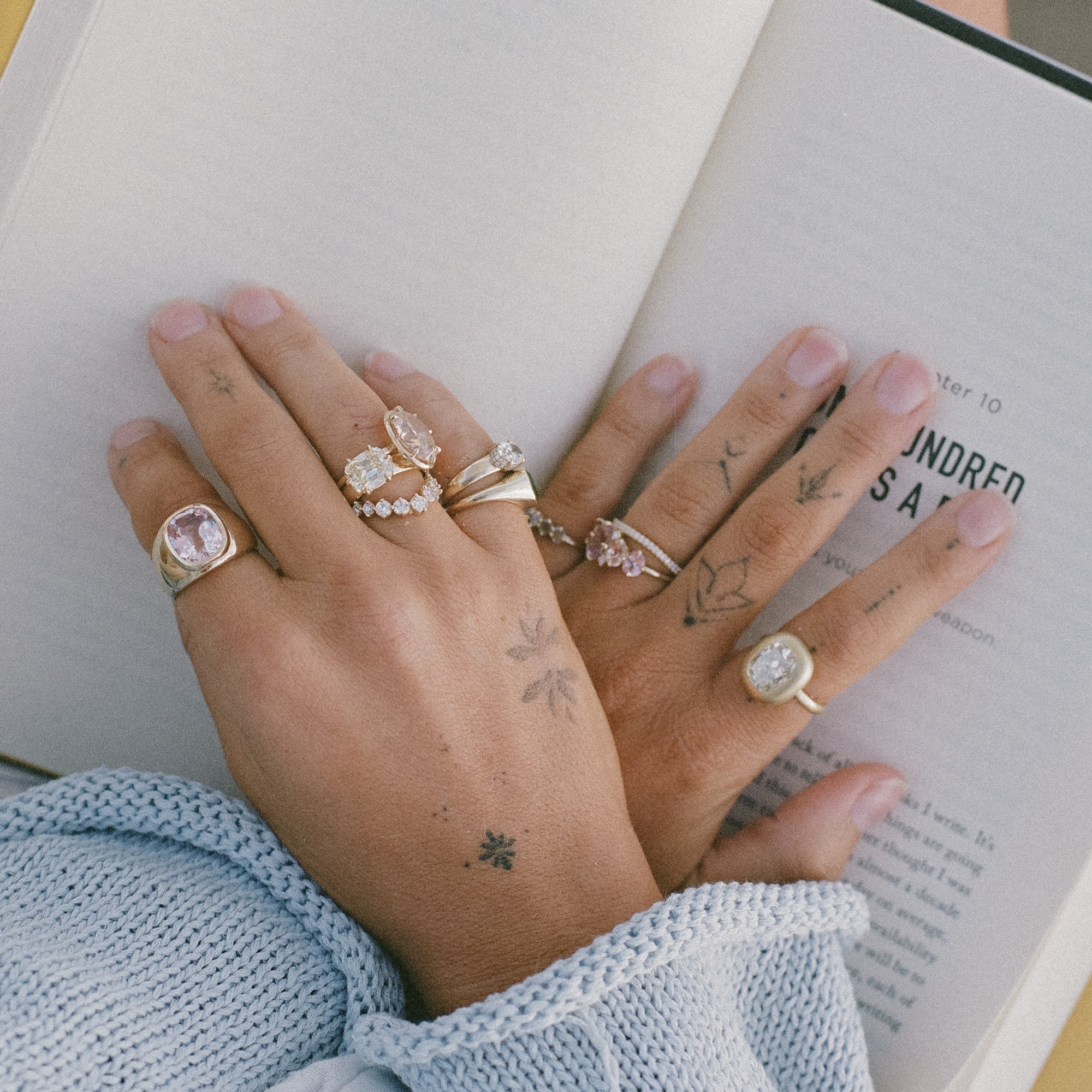 Womans Tattooed Hands Crossed On Open Book Pages With A Variety Of One Of A Kind Diamond And Gemstone Engagement Rings