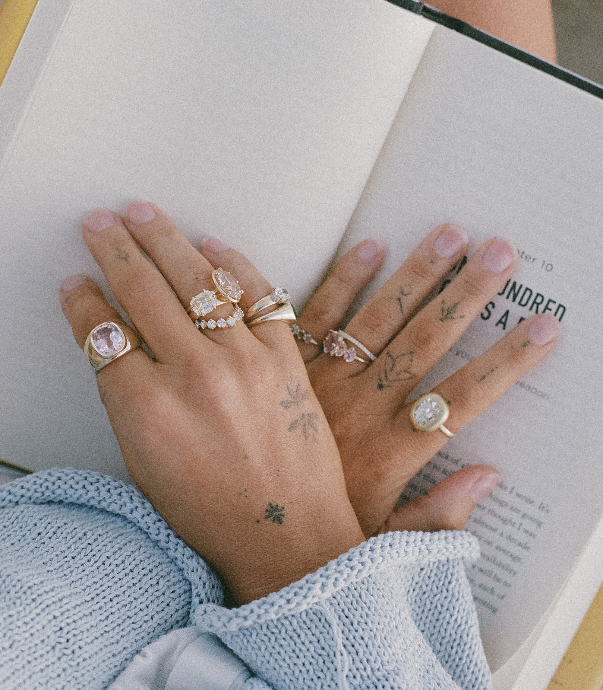 Womans Tattooed Hands Crossed On Open Book Pages With A Variety Of One Of A Kind Diamond And Gemstone Engagement Rings
